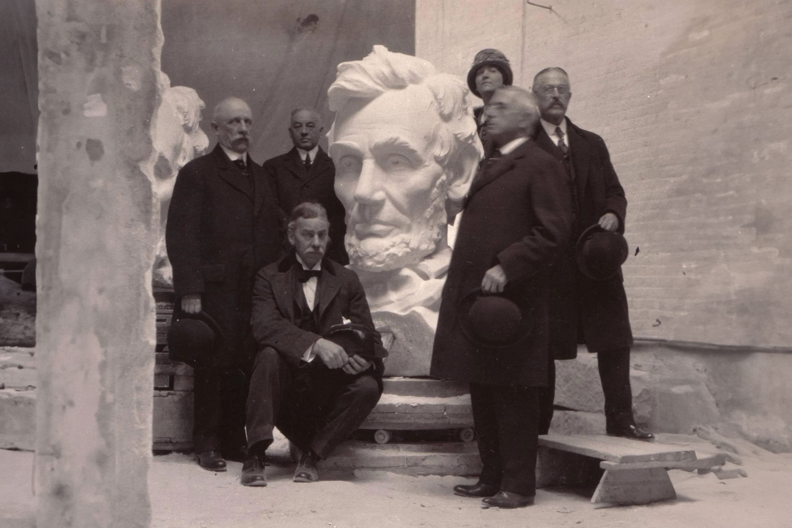 A historic black-and-white photo of a group of men and one woman, Evelyn Beatrice Longman, standing around the head of the Lincoln Memorial statue, showcasing the artists and craftsmen who worked on the iconic monument.